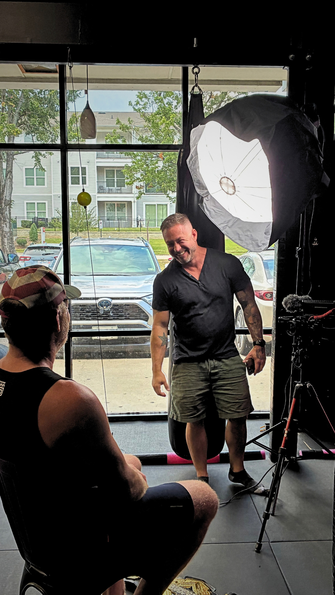 Brad working with a seated interview subject beside a large softbox light and camera on a tripod during an on-site shoot